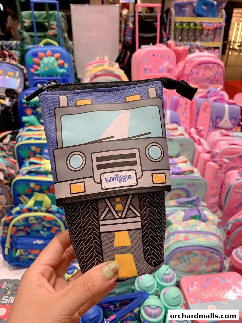 A hand holds a Smiggle pencil case shaped like a truck, with colorful backpacks in the background.
