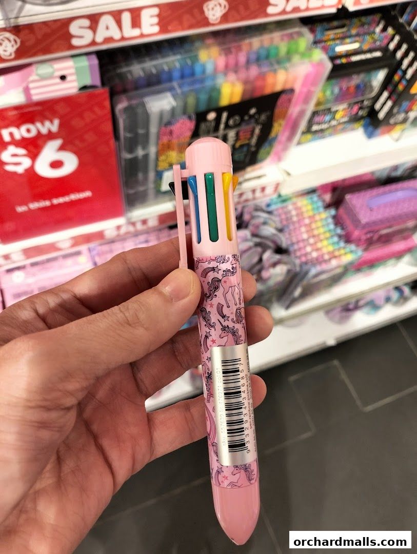 A hand holds a pink multi-color pen with unicorn designs, with sale signs and stationery in the background.