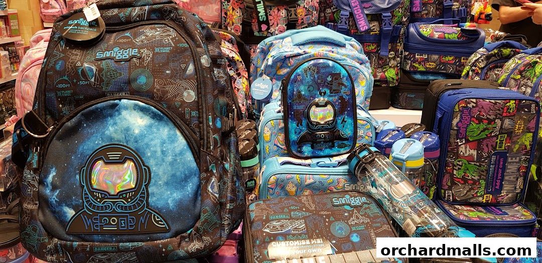 A vibrant display of Smiggle backpacks, lunchboxes, and water bottles with space and dinosaur themes.