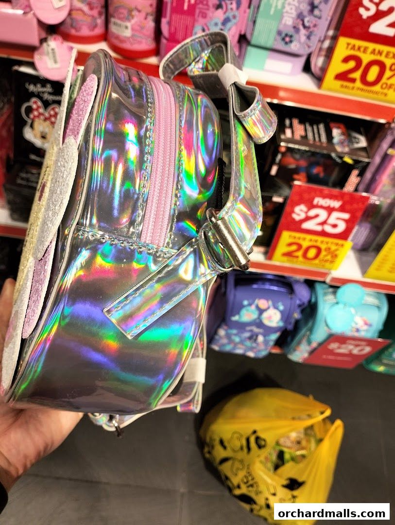 A hand holds a shiny holographic backpack with pink accents in a store.