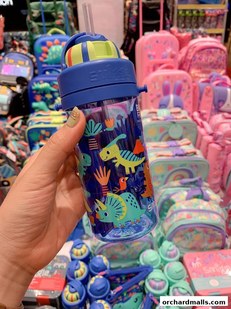 A hand holds a dinosaur-themed Smiggle water bottle with colorful backpacks in the background.