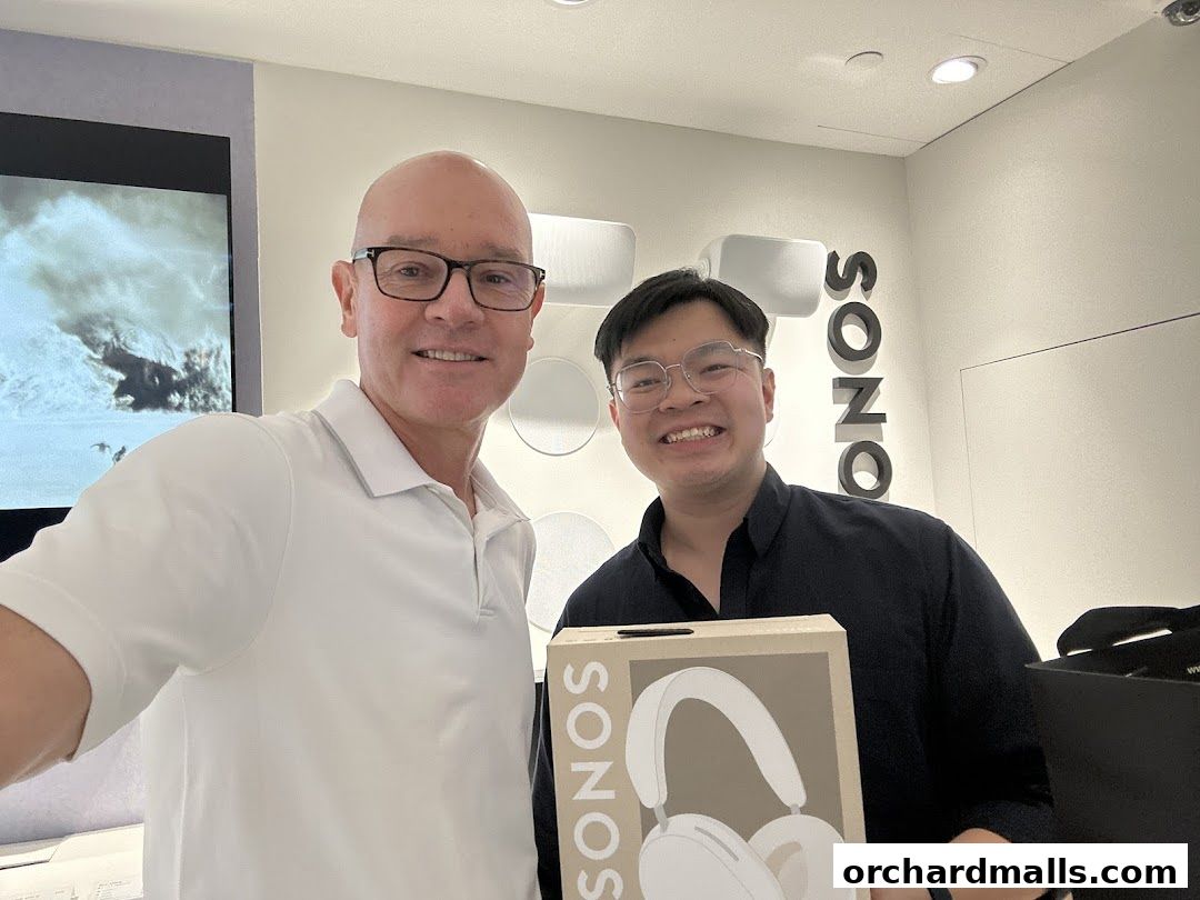 Two smiling men hold a Sonos headphones box inside a modern retail store.