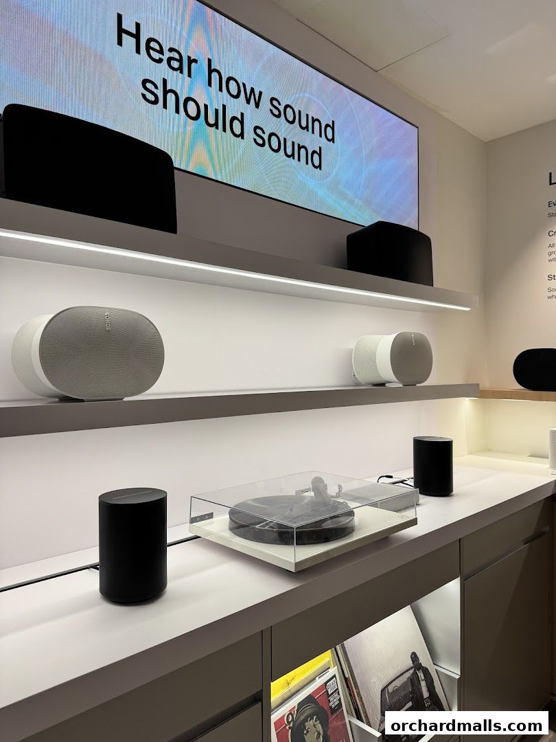Sonos speakers and a turntable displayed on shelves with a large screen showing 'Hear how sound should sound'.