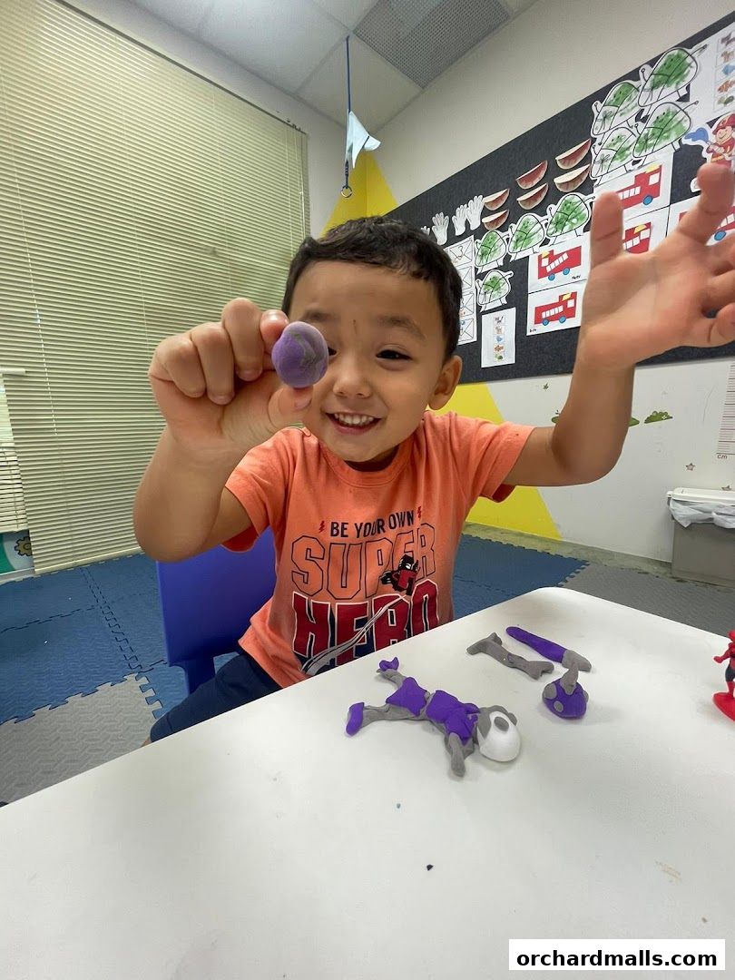 A smiling child holds up a purple clay ball, with clay figures on a table.