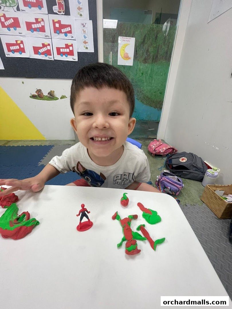 A smiling child plays with colorful modeling clay at a table in a classroom.