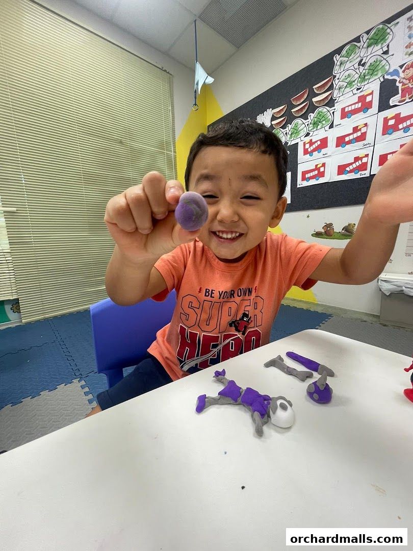 A smiling child holds up a purple clay ball, with clay figures on the table.