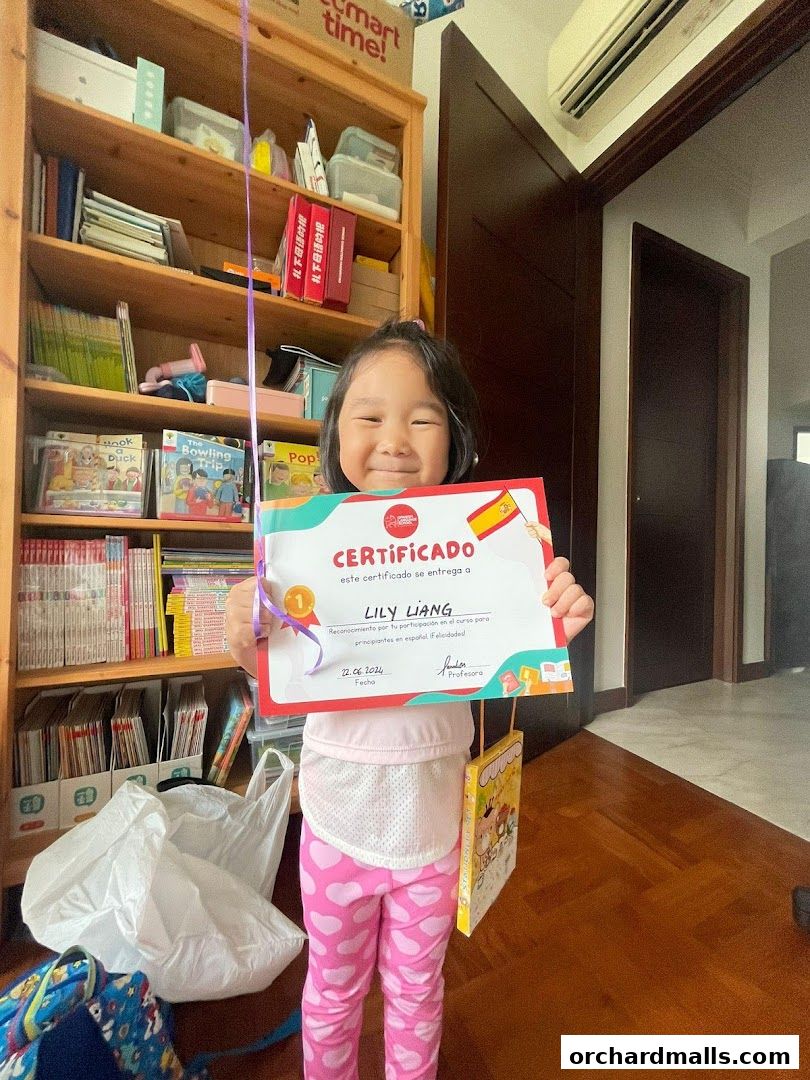 A young girl proudly holds a Spanish language certificate in front of a bookshelf.