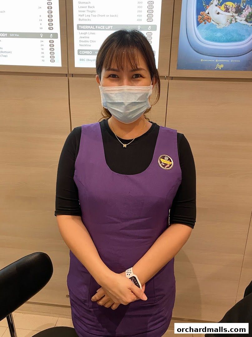 A woman wearing a mask and a purple uniform stands in front of a price list.