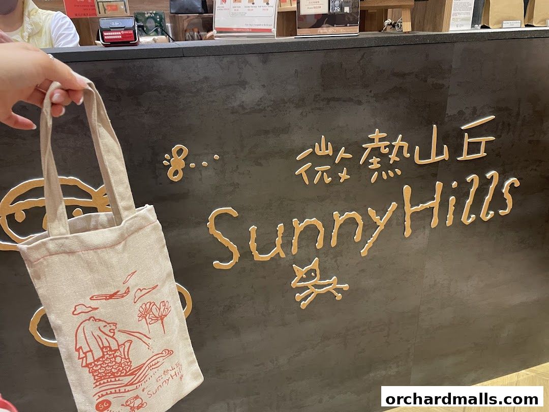 A hand holds a tote bag in front of the SunnyHills Orchard shop logo.