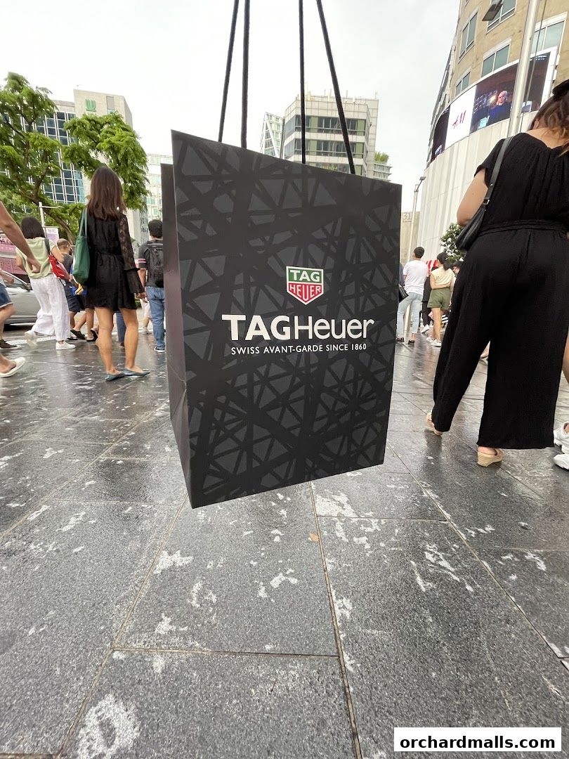 A large TAG Heuer shopping bag is displayed outdoors with people walking by.