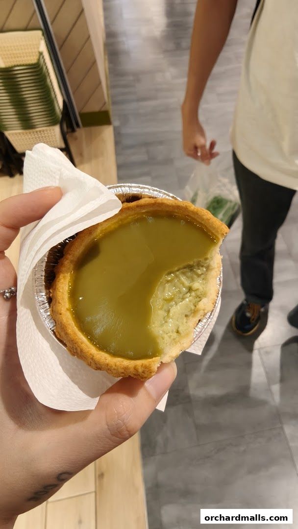 A hand holds a partially eaten green egg tart from Tai Cheong Bakery.