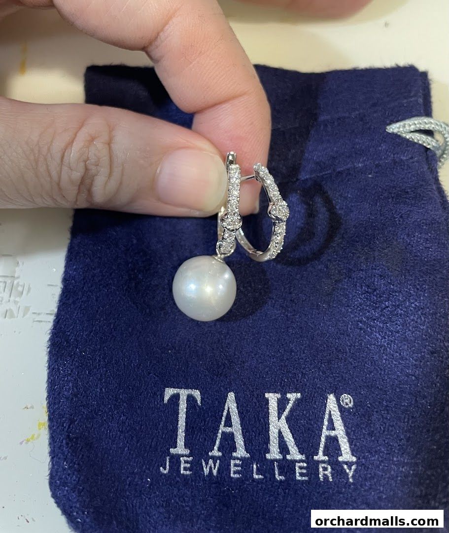 A hand holds a pearl and diamond earring over a blue pouch with the TAKA Jewellery logo.