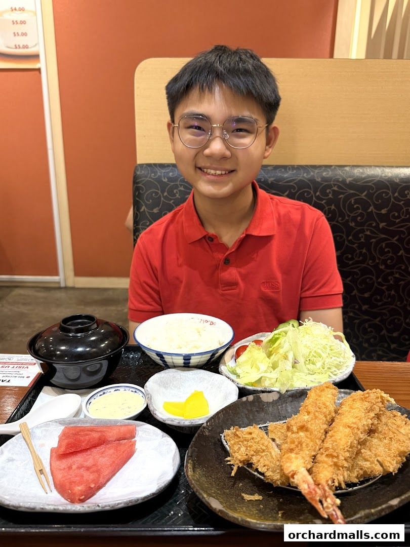 A young person smiles at the camera with a meal of fried seafood, rice, salad, and watermelon.