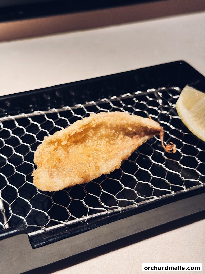 A close-up of a golden-brown tempura piece served with a lemon wedge on a wire rack.