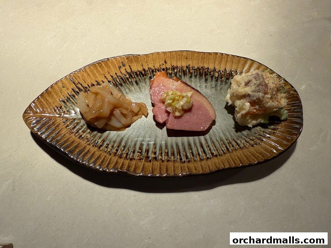 A leaf-shaped plate holds three small appetizers: a seafood salad, sliced duck, and potato salad.