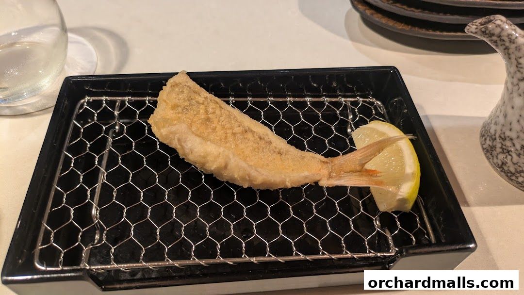 A perfectly fried tempura fish served with a slice of lemon on a black wire rack.