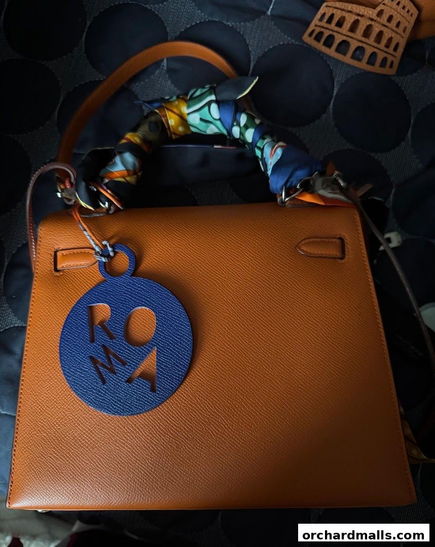An orange leather handbag with a colorful scarf tied around the handle and a 'ROMA' charm.