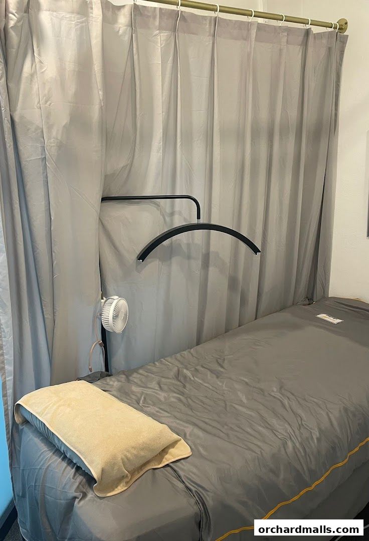 A treatment bed with a pillow, fan, and overhead light in a room with grey curtains.