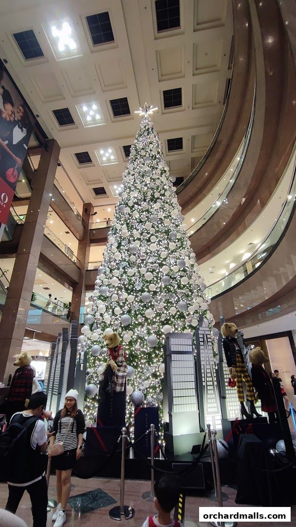 A large, decorated Christmas tree stands in a mall atrium with people posing for photos.
