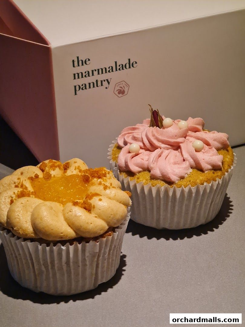 Two beautifully decorated cupcakes with frosting and toppings, next to a branded box.