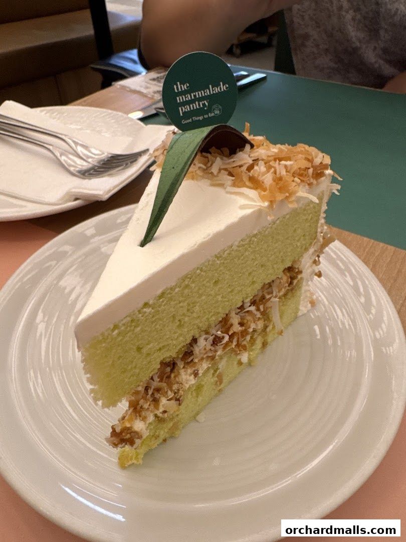 A slice of green pandan cake with coconut flakes and cream on a white plate.