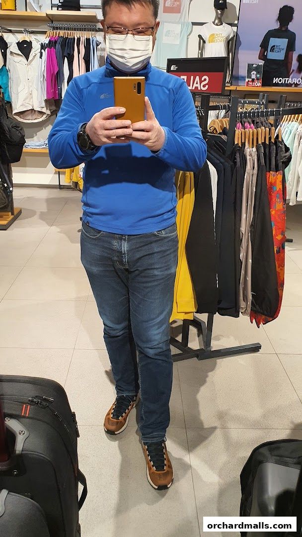 Man in a blue jacket and jeans takes a selfie in a store with racks of clothing.