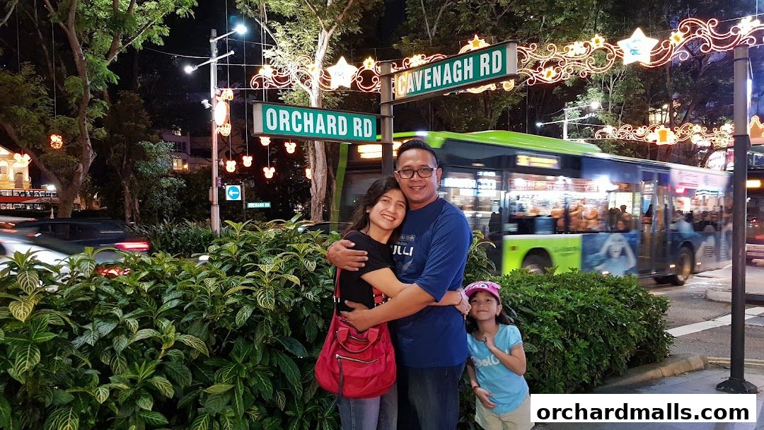 A family poses for a photo on Orchard Road at night, with festive lights and a bus in the background.