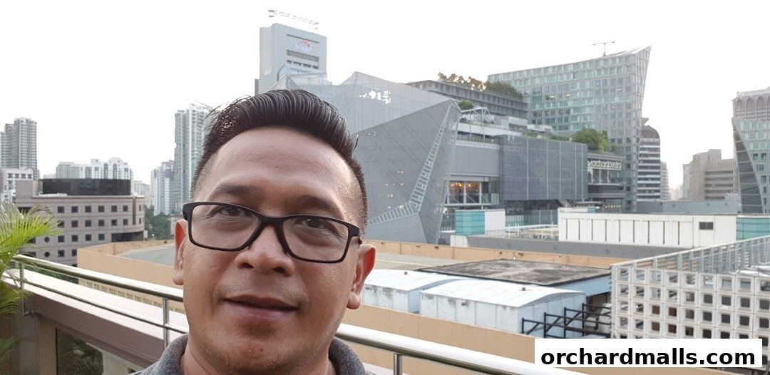 A man wearing glasses takes a selfie with a modern cityscape in the background.