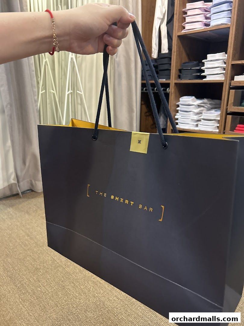 A hand holds a shopping bag from 'The Shirt Bar' with shirts displayed in the background.