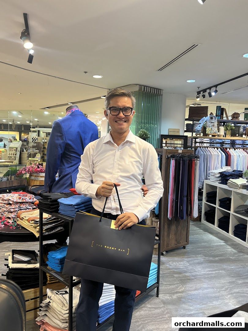 A man holds a shopping bag from The Shirt Bar inside a well-stocked clothing store.
