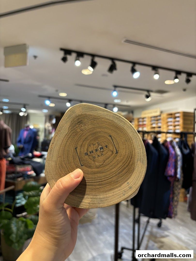 A hand holds a wooden coaster with 'The Shirt Bar' logo inside a clothing store.