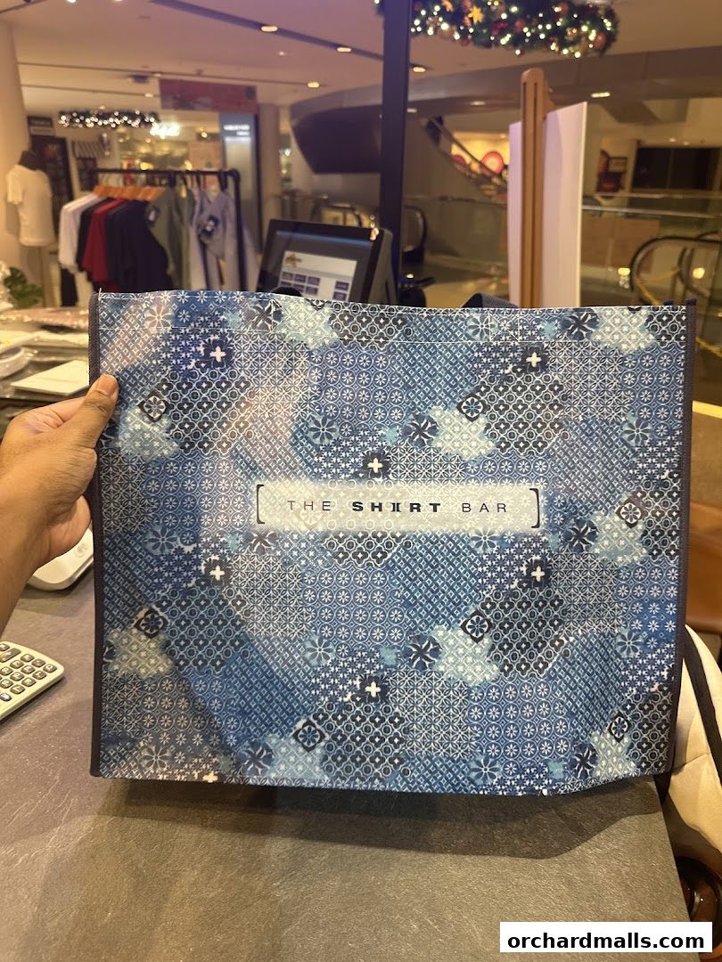 A person holds a patterned shopping bag from The Shirt Bar in a retail store.
