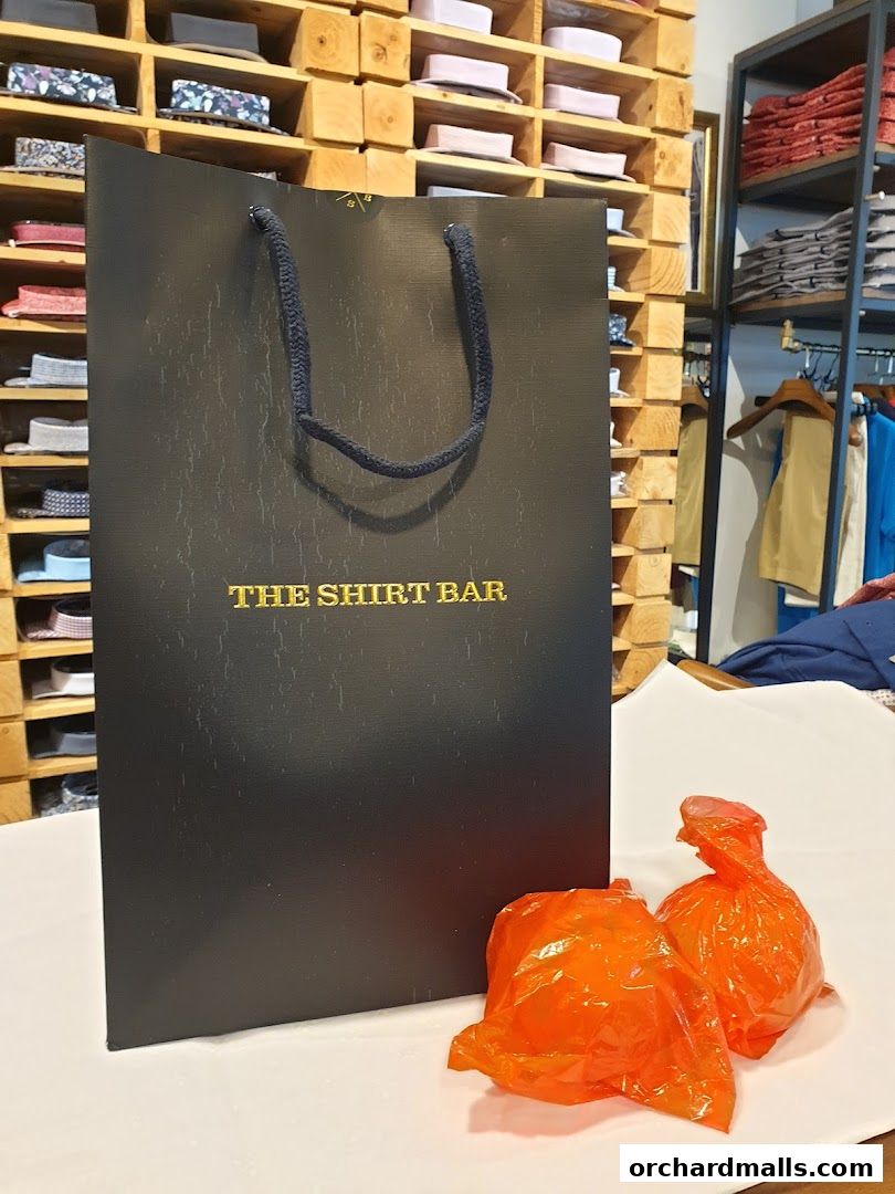 A black shopping bag with 'The Shirt Bar' logo and two orange bags on a table.