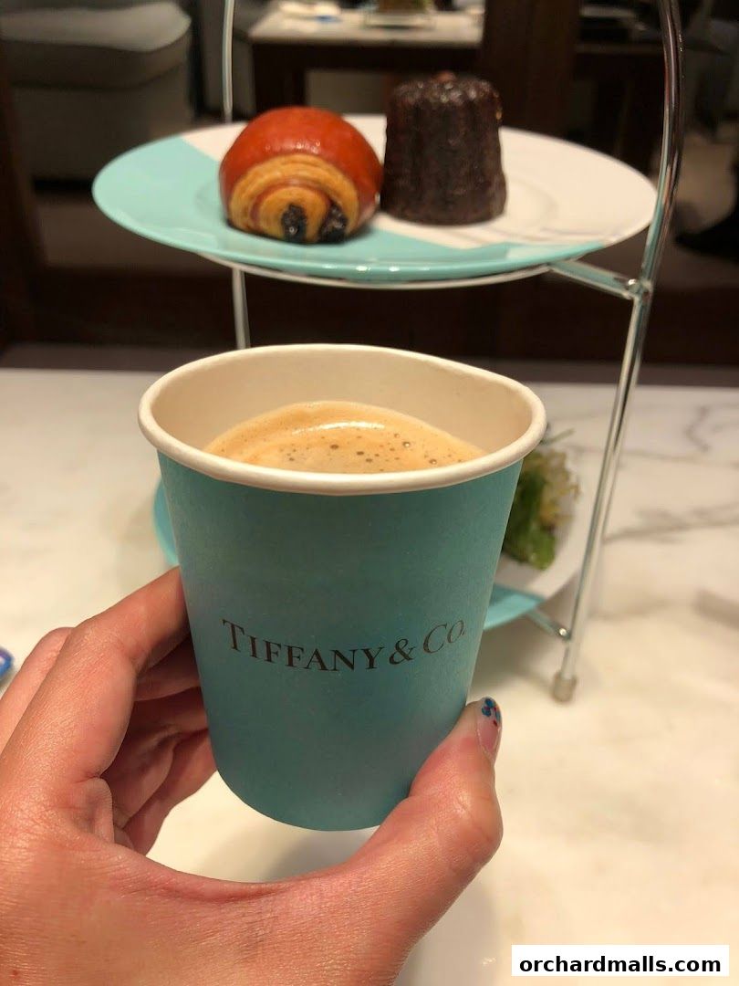 A hand holds a Tiffany & Co. branded cup of coffee with pastries on a tiered stand.