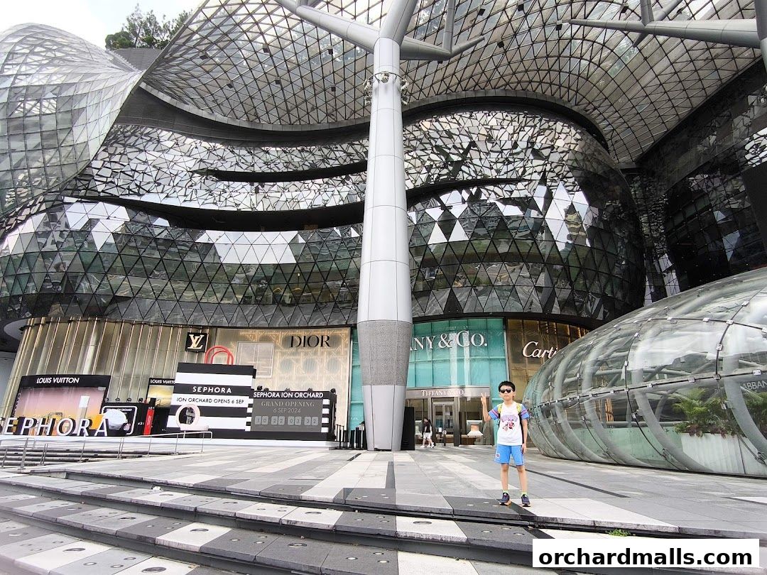 A person stands in front of a modern building with multiple luxury brand stores.