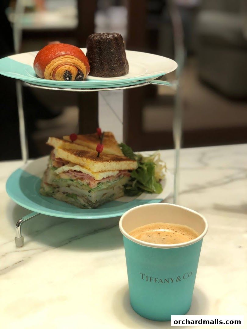 A tiered stand holds a pastry and a sandwich, with a Tiffany & Co. branded coffee cup in the foreground.