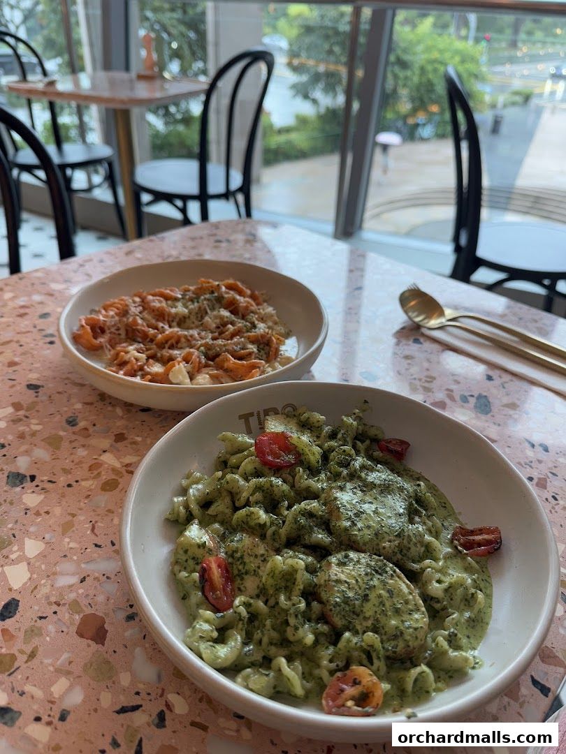 Two bowls of pasta dishes served on a terrazzo table with chairs and window view.