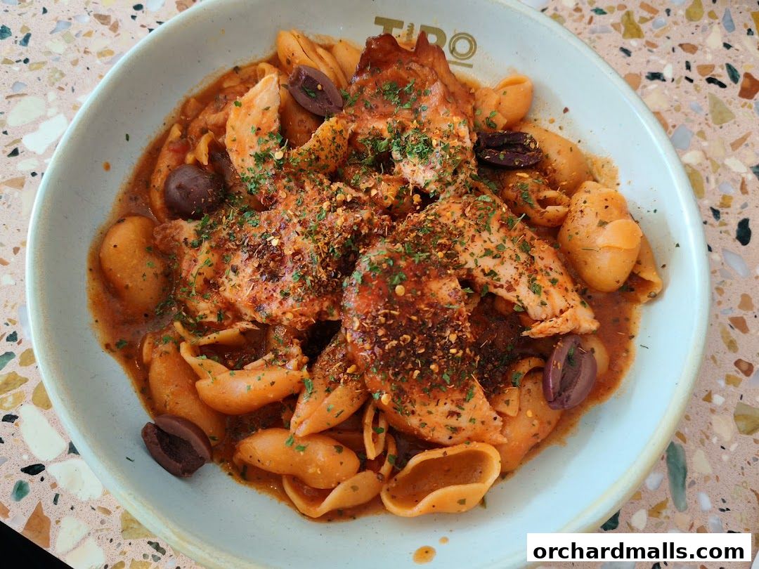 A close-up of a pasta dish with chicken and olives, seasoned with herbs and chili flakes.