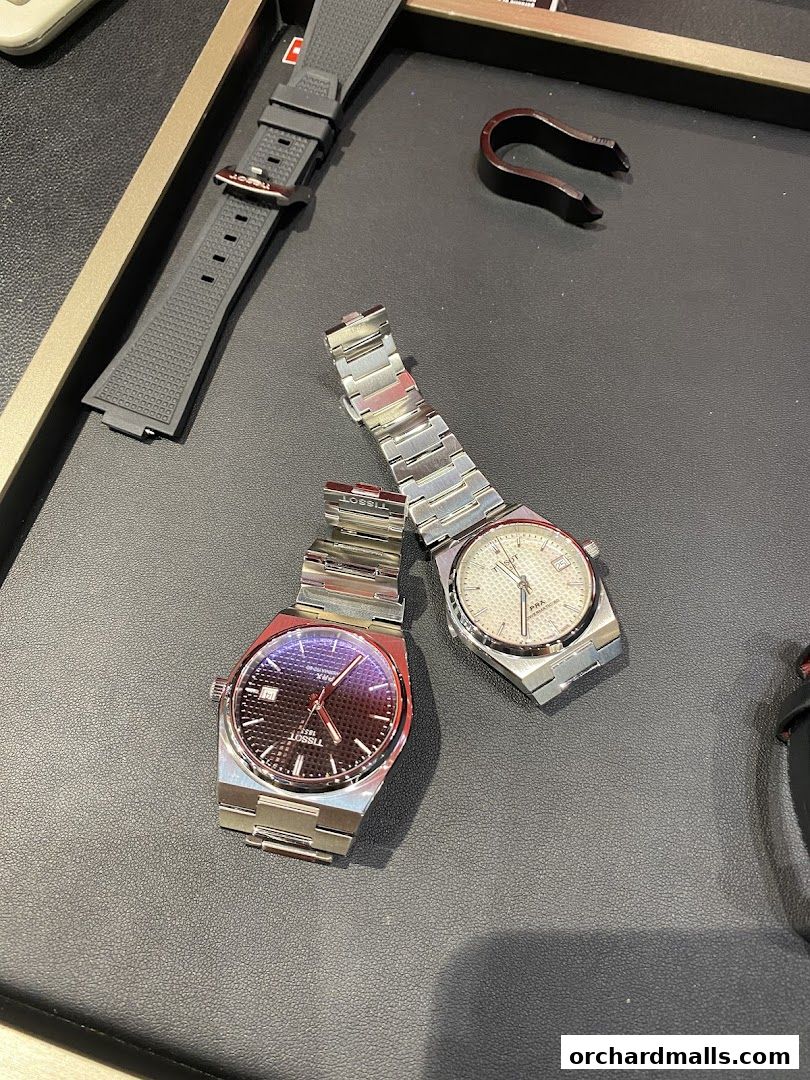 Two Tissot watches, one with a dark dial and one with a white dial, displayed on a dark surface.