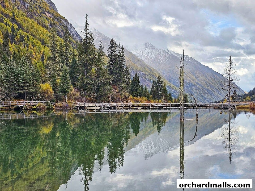 A serene lake reflects a forest and snow-capped mountains under a cloudy sky.