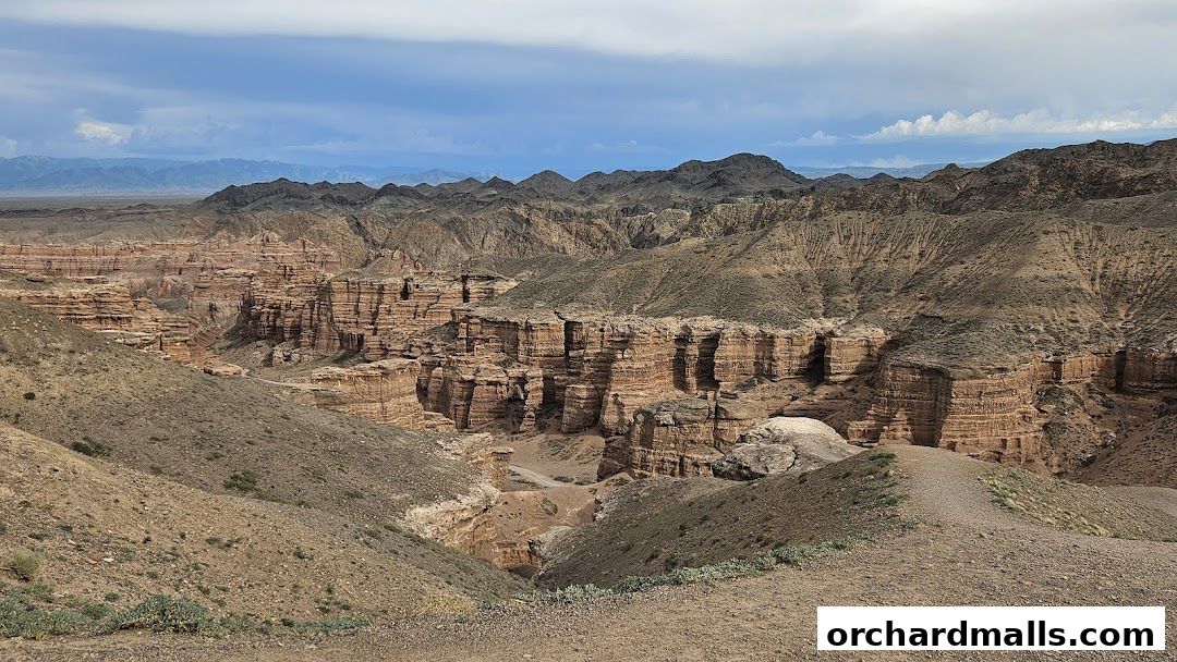 A vast, rugged canyon landscape with layered rock formations under a cloudy sky.