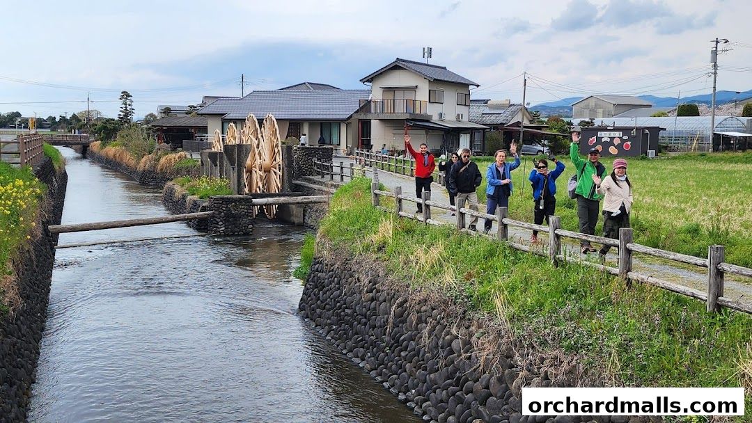 Tourists wave while walking along a canal with water wheels and traditional houses.