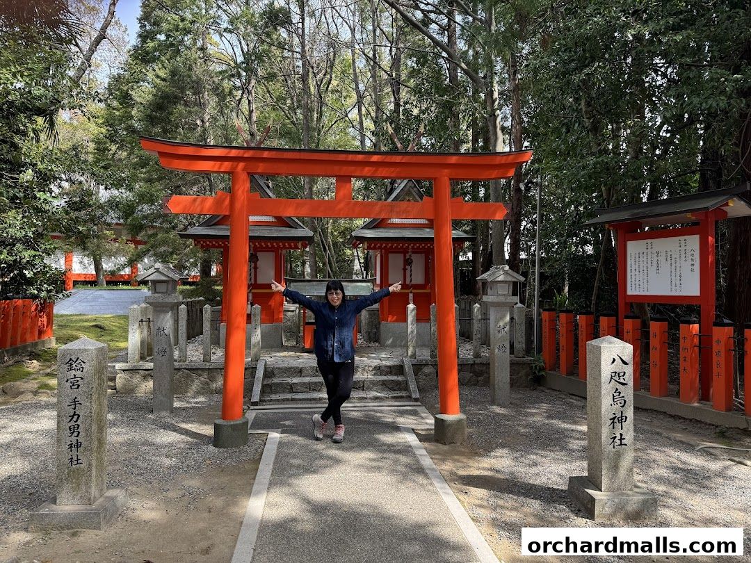 A person stands in front of a vibrant orange torii gate at a Japanese shrine.