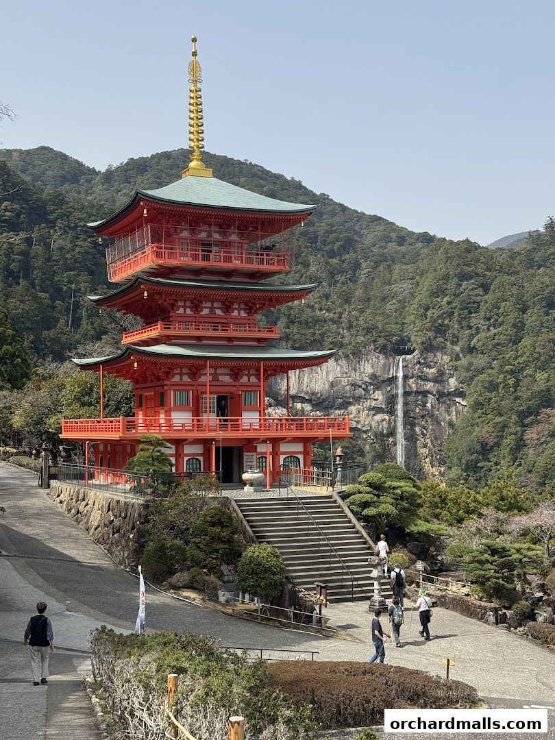 A vibrant red pagoda stands tall against a backdrop of lush green mountains and a cascading waterfall.