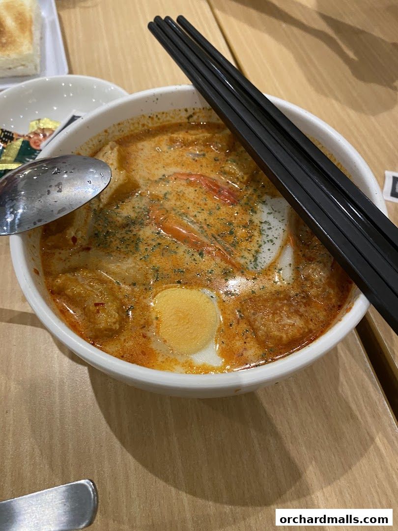 A close-up of a bowl of Laksa with a spoon and chopsticks on a wooden table.