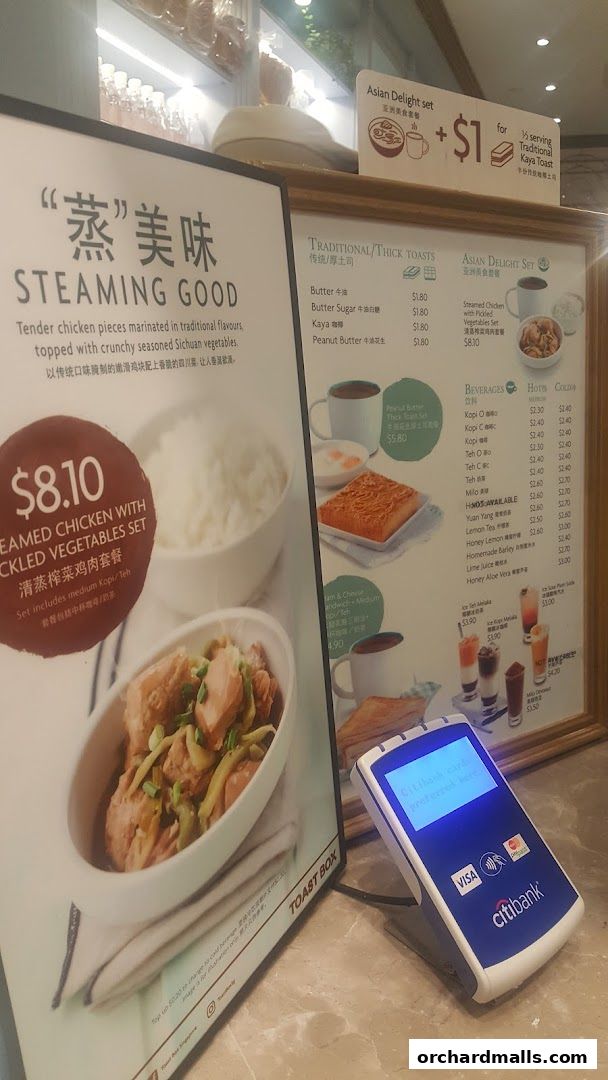 A menu board displaying various food and beverage options from Toast Box, with a payment terminal in the foreground.