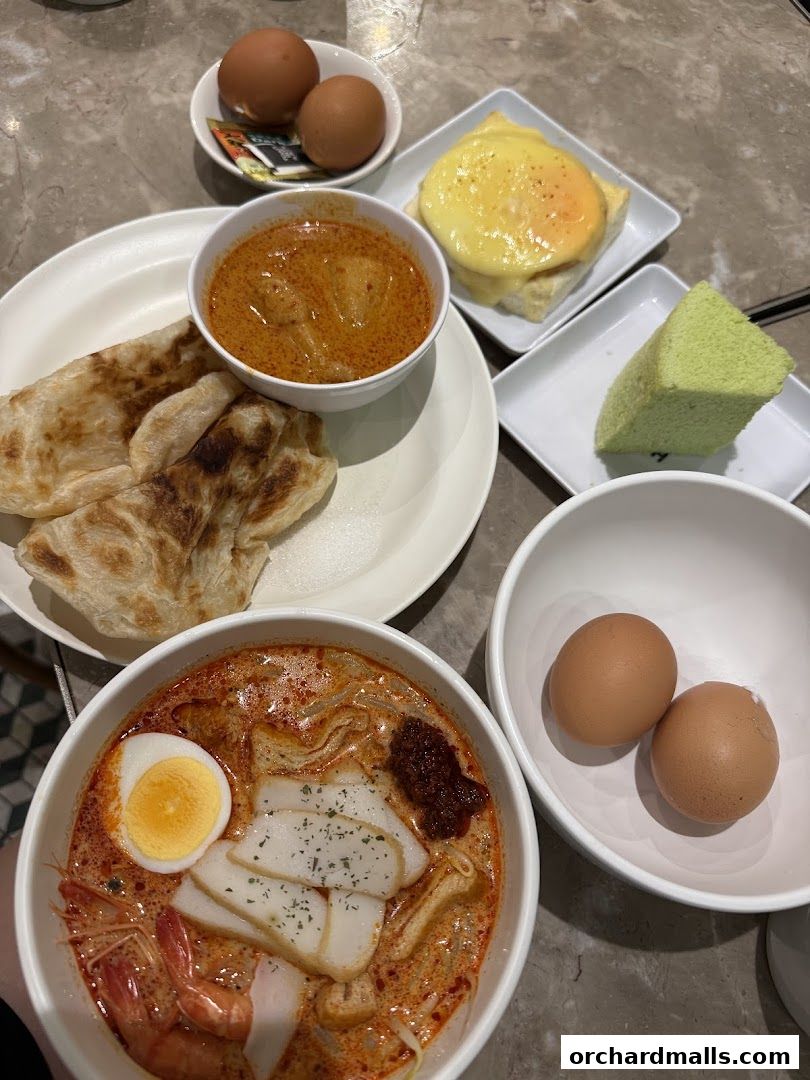 A spread of delicious Southeast Asian breakfast dishes including laksa, curry, roti, eggs, and cake.
