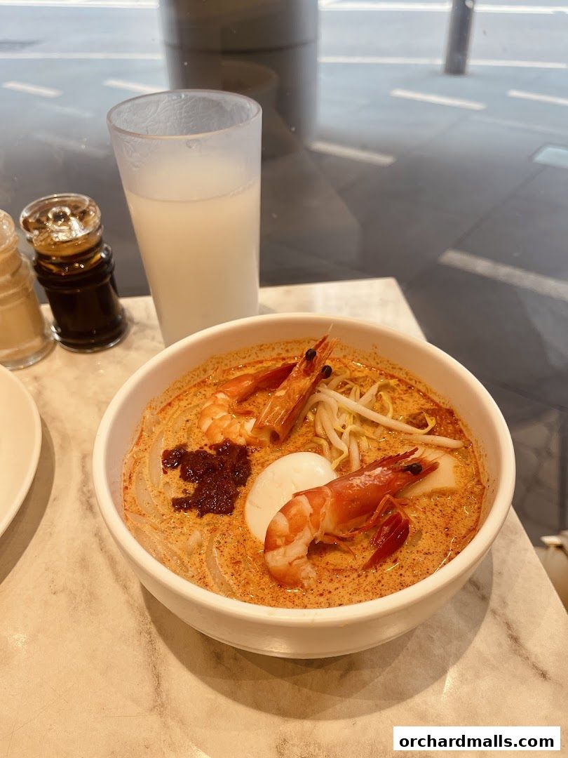 A bowl of Laksa with prawns, egg, and bean sprouts, served with a drink.