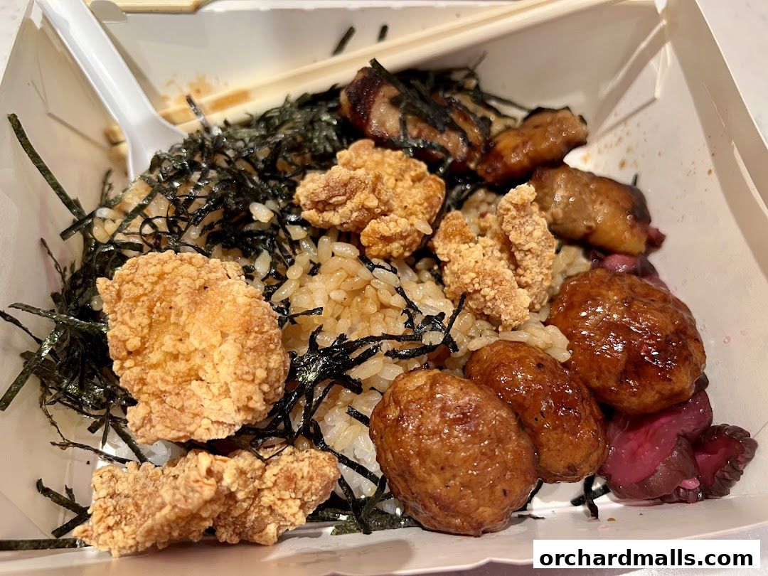 A close-up of a takeaway box filled with rice, fried chicken, meatballs, and seaweed.