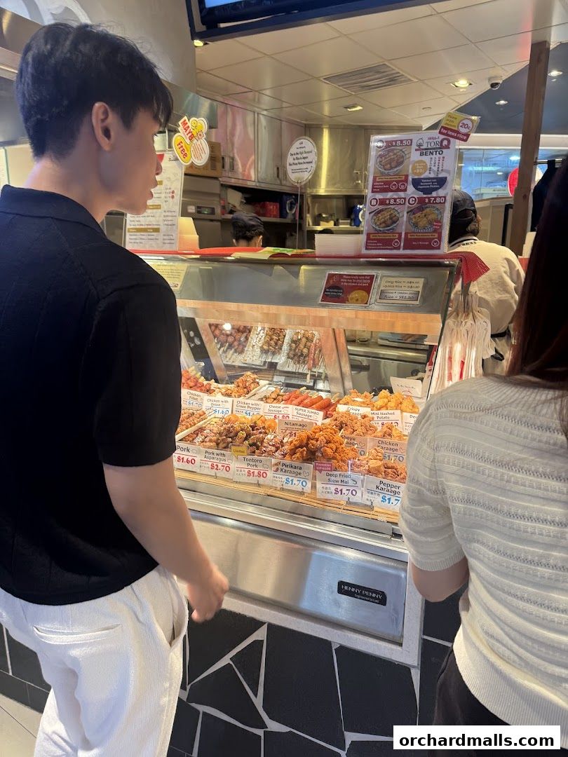 Customers view a display of various grilled skewers and fried items at a food stall.